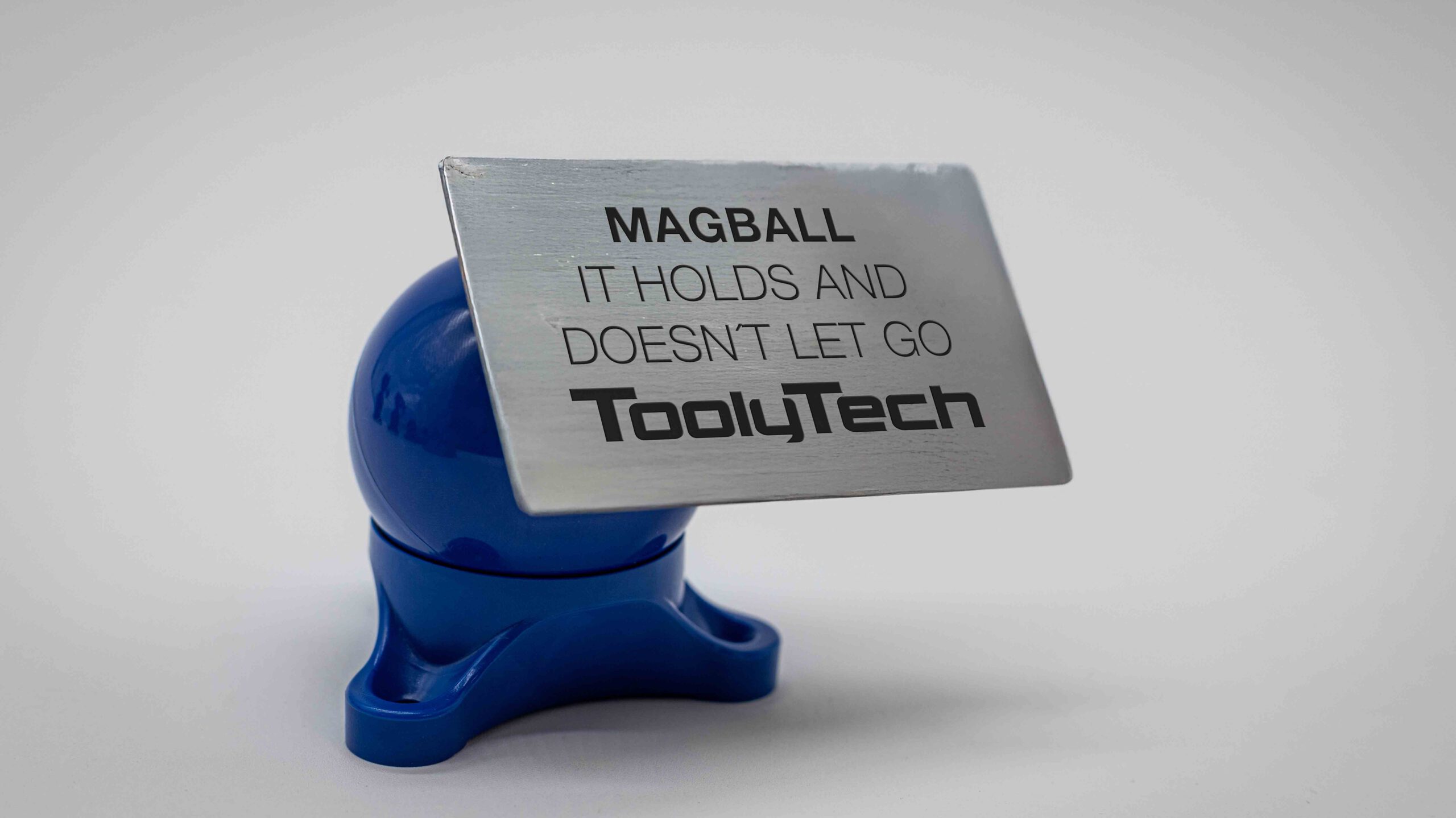Die blaue Magnetkugel hält eine gebürstete Metallkarte mit dem Text „MAGBALL“ und „IT HOLDS AND DOESN'T LET GO“.