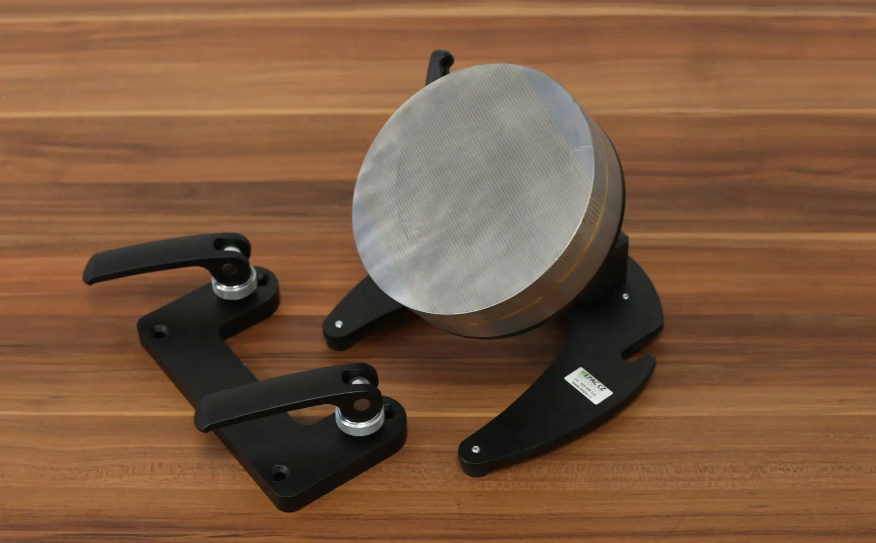 Ein schwarzer Magnetspannhalter hält ein rundes Metallwerkstück neben zwei Befestigungsklemmen auf einer Holzoberfläche.