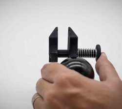 Eine Hand hält eine schwarze Federklemme mit Metallfeder vor hellem Hintergrund, während der Daumen den Mechanismus seitlich festdrückt.