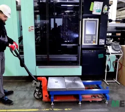 Ein Arbeiter mit Helm und roten Handschuhen rangiert einen mobilen Werkzeugöffner neben einer Spritzgussmaschine in der Produktionshalle.