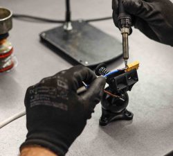 Mit schwarzen Handschuhen wird eine kleine elektrische Verbindung mit dem Lötkolben an einem Bauteil auf dem Tisch verlötet.