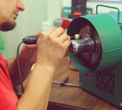 Ein Mann in rotem Shirt bearbeitet einen Metallzylinder an einer grünen Poliermaschine mit einem kleinen Schleifaufsatz.