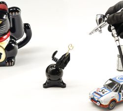Schwarze Maneki-Neko-Figur, Airbrush-Pistole mit Handschuh und weißes Modellauto mit Startnummer 54 stehen auf einem hellen Tisch.
